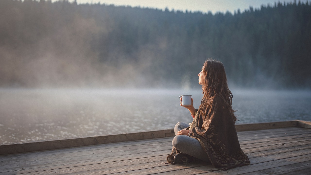 Frau sitzt mit einer Tasse auf einem Steg am nebligen See