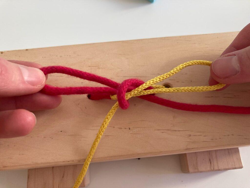 Masche binden Schritt 6: Fertige Masche aus roter und gelber Schnur am Übungsbrett
