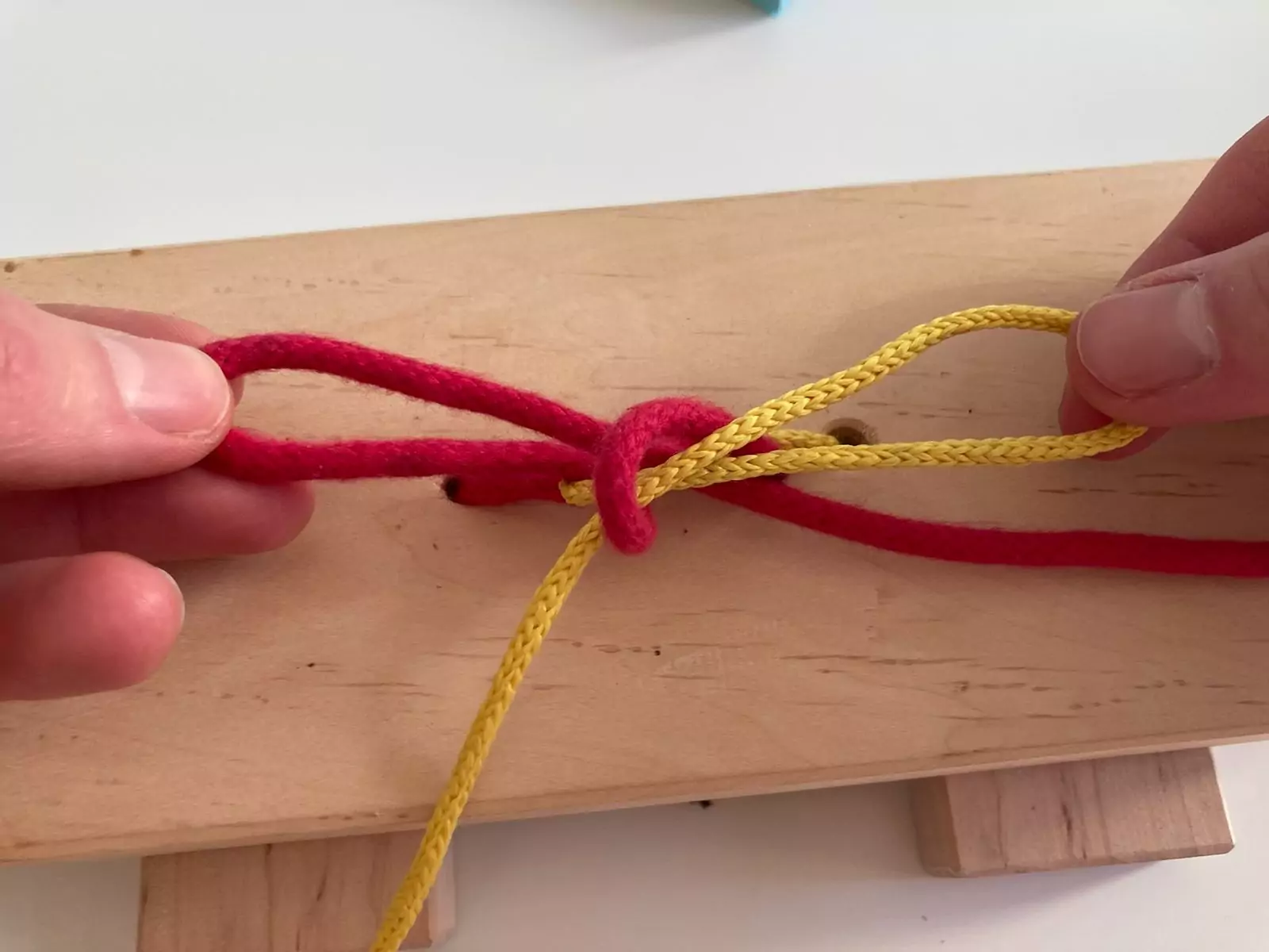 Hände üben an einem Holzbrett mit roter und gelber Schnur das Maschebinden