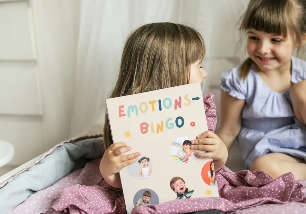 Zwei Mädchen lachen, eines hält das Emotions-Bingo mit Fotos von Kindergesichtern