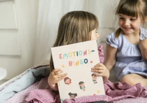 Zwei Mädchen lachen, eines hält das Emotions-Bingo mit gezeichneten Gefühlsfiguren