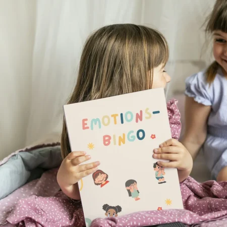 Zwei Mädchen lachen, eines hält das Emotions-Bingo mit gezeichneten Gefühlsfiguren
