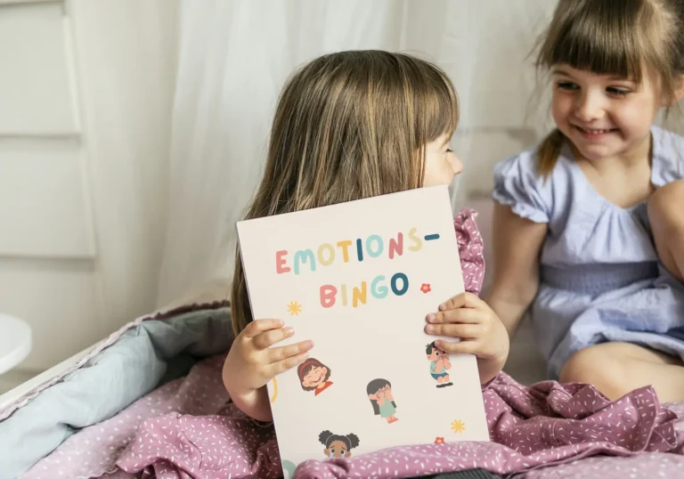 Zwei Mädchen lachen, eines hält das Emotions-Bingo mit gezeichneten Gefühlsfiguren