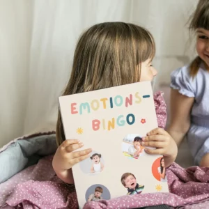 Zwei Mädchen lachen, eines hält das Emotions-Bingo mit Fotos von Kindergesichtern