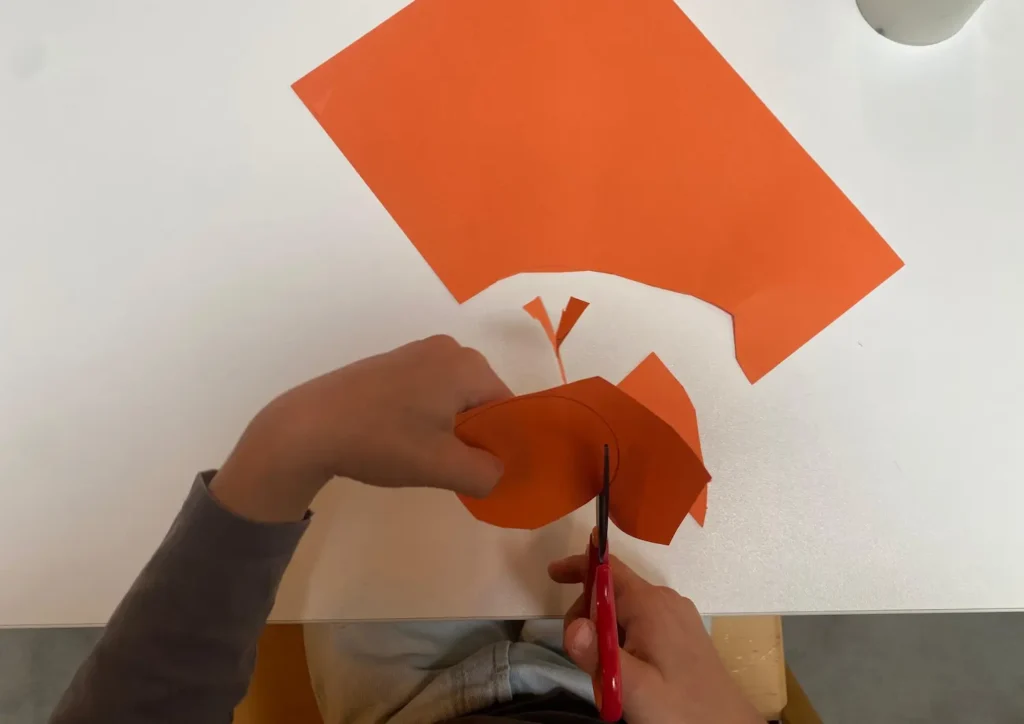 Kind schneidet mit einer roten Schere ein Stück orangefarbenes Papier