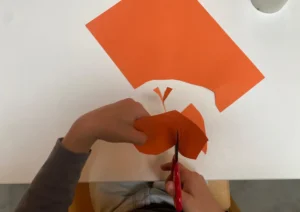 Kind schneidet mit einer roten Schere ein Stück orangefarbenes Papier