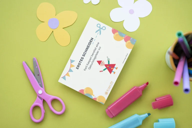 Erstes Schneiden 1: Titelseite der Schneidegeschichte neben Kinderschere, Buntstiften und Papierblumen